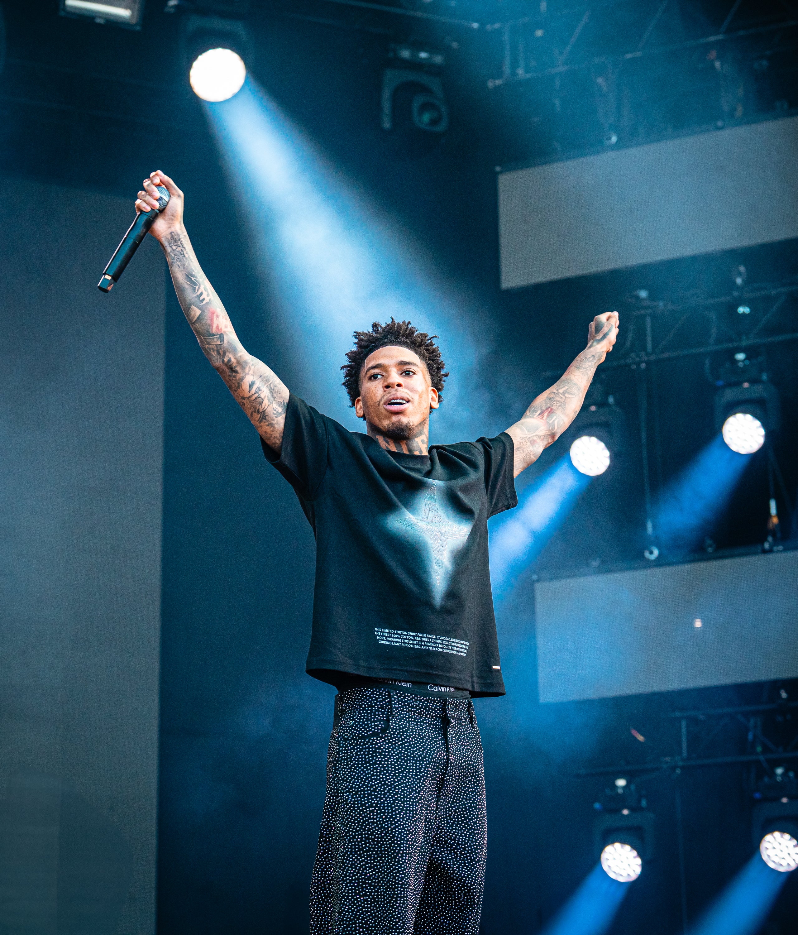 NLE Choppa on stage wearing Finelli Star T-Shirt with arms raised, holding a microphone, under bright stage lights.